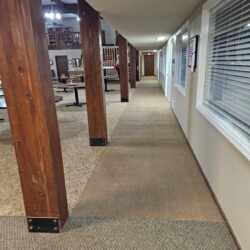 Long hallway with wooden support beams, carpeted floor, and windows and doors along the right side.