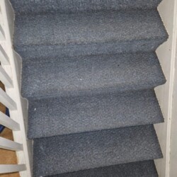 A set of steep, narrow stairs covered with blue-grey carpet, seen from above. White railings are on the left side.