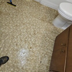 A bathroom floor with tile pattern is wet, showing water pooling near a toilet and a broom leaning against the wall.